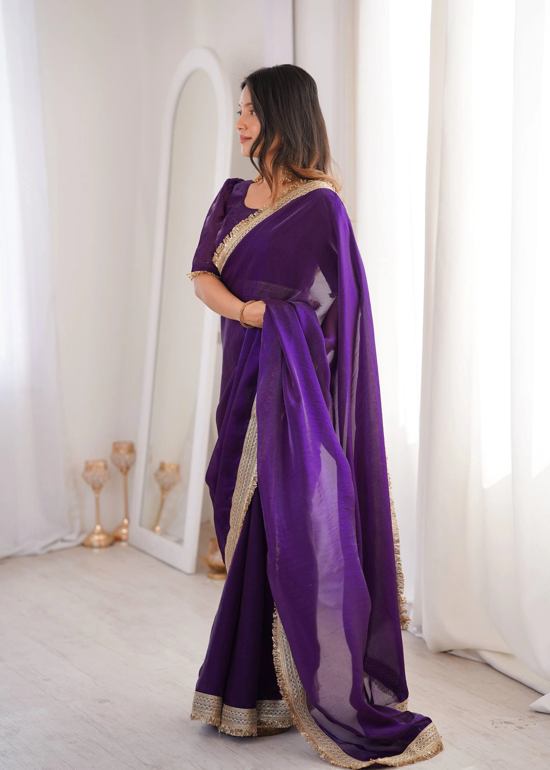 Woman wearing a purple saree with gold border in a room with white curtains.